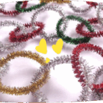 Colorful metallic pipe cleaners twisted into rings and spirals are scattered on a white surface, alongside two small yellow decorative pieces.
